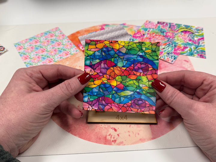 A person holding a 4x4 inch acrylic stained glass craft piece with a multicolored floral pattern.