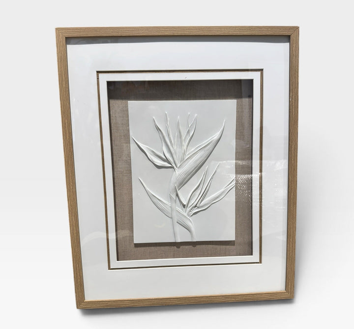 Framed botanical wall art featuring a white embossed leaf design on a light gray background, surrounded by white matting and a natural wood frame, photographed against a clean white wall.