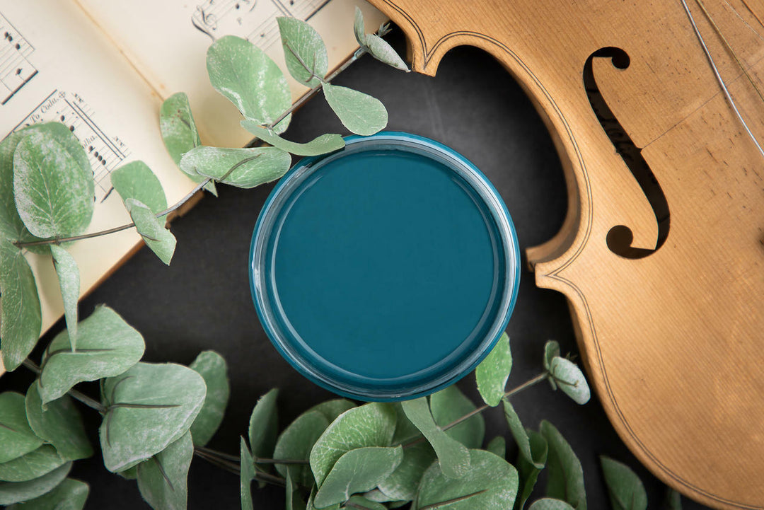 Open container of Antebellum Blue Chalk Mineral Paint against a black background with a cello